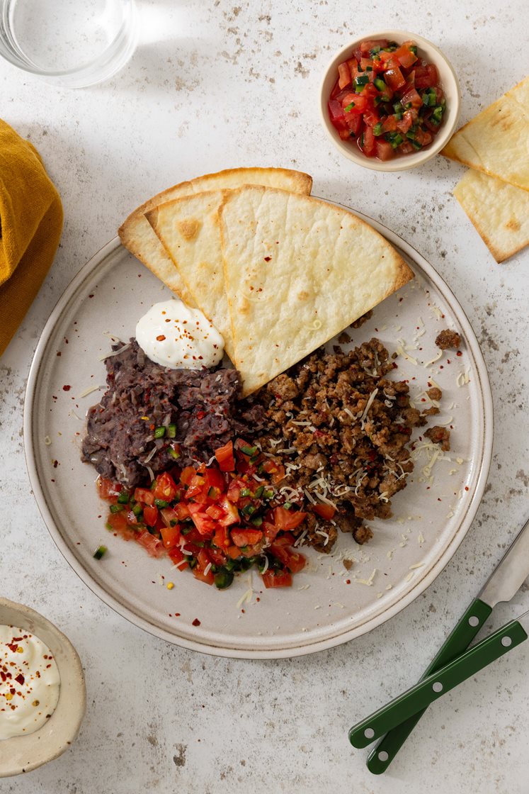 Tostada med refried beans, kjøttdeig med grønnsaker toppet med revet ost, rømme og jalapeno